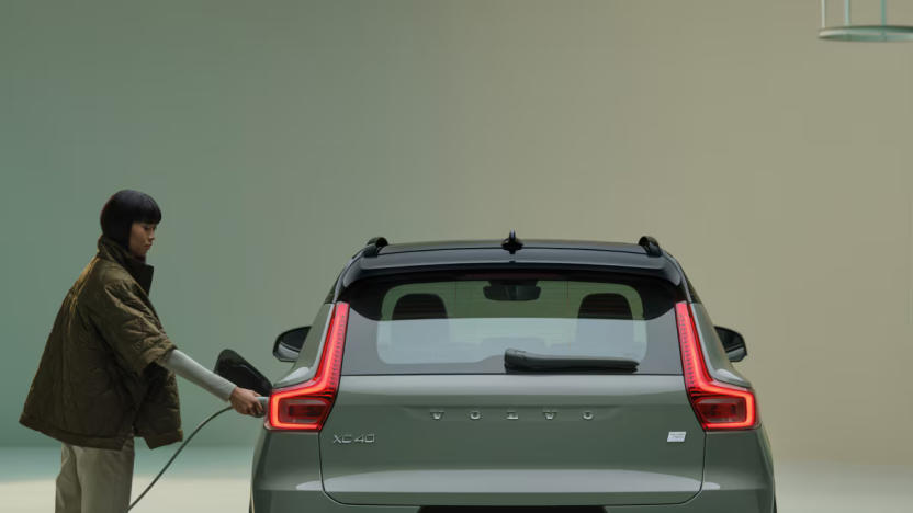 A person holding a charger up to the left side of a Volvo EV. Rear view of vehicle.