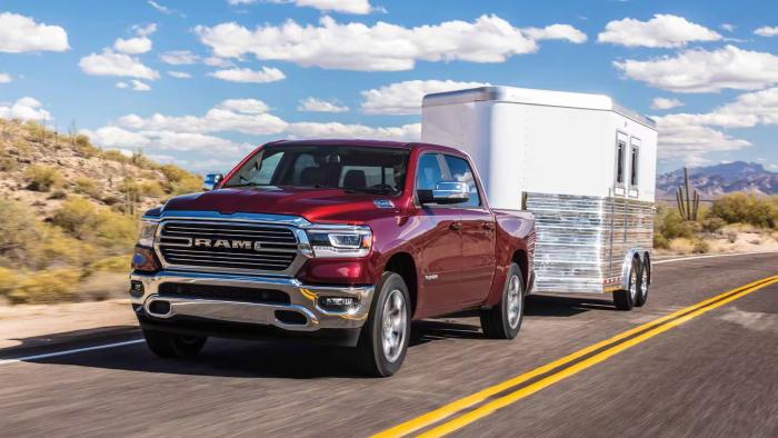 A red Ram 1500 truck towing a small trailer on a highway with a desert seen in the background and cloudy blue sky