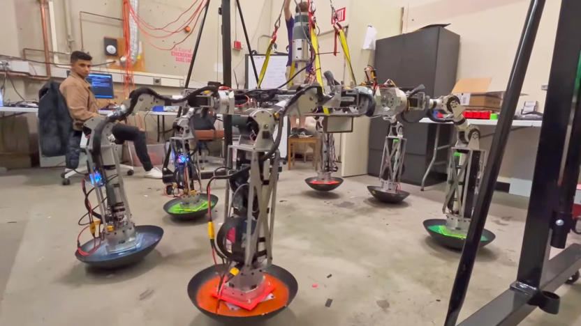 Photo inside an MIT lab of a student looking over a spider-like robot with six worm-like legs and wok-shaped “shoes.”