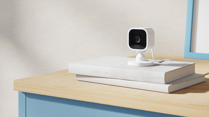 The black and white Blink Mini security camera sits on a pile of books on a wooden cabinet. 